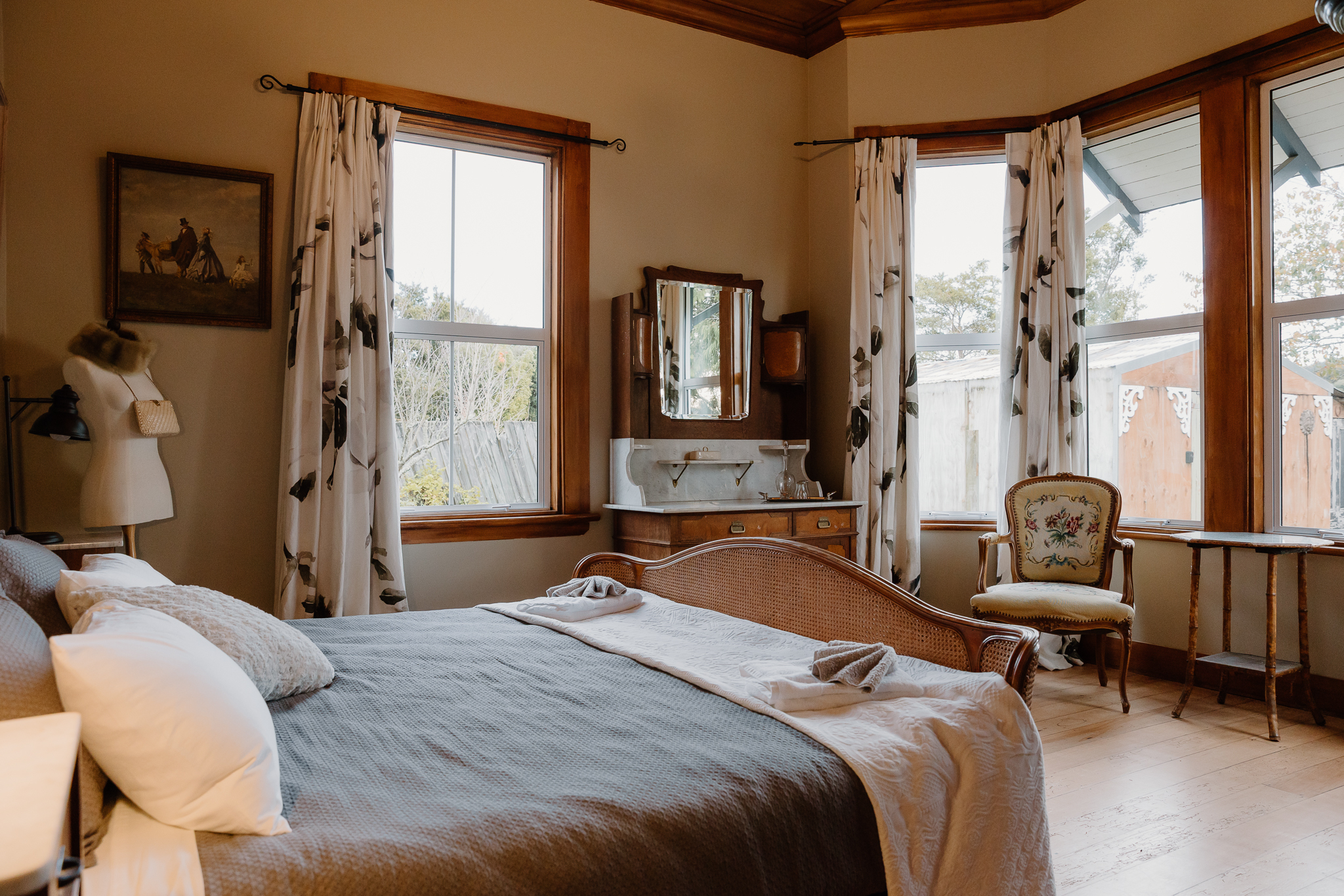 Main guest bedroom at Willow Bank Villa with vintage decor, featuring a cane vintage bed, floral armchair, antique vanity, and large bay windows with garden views.