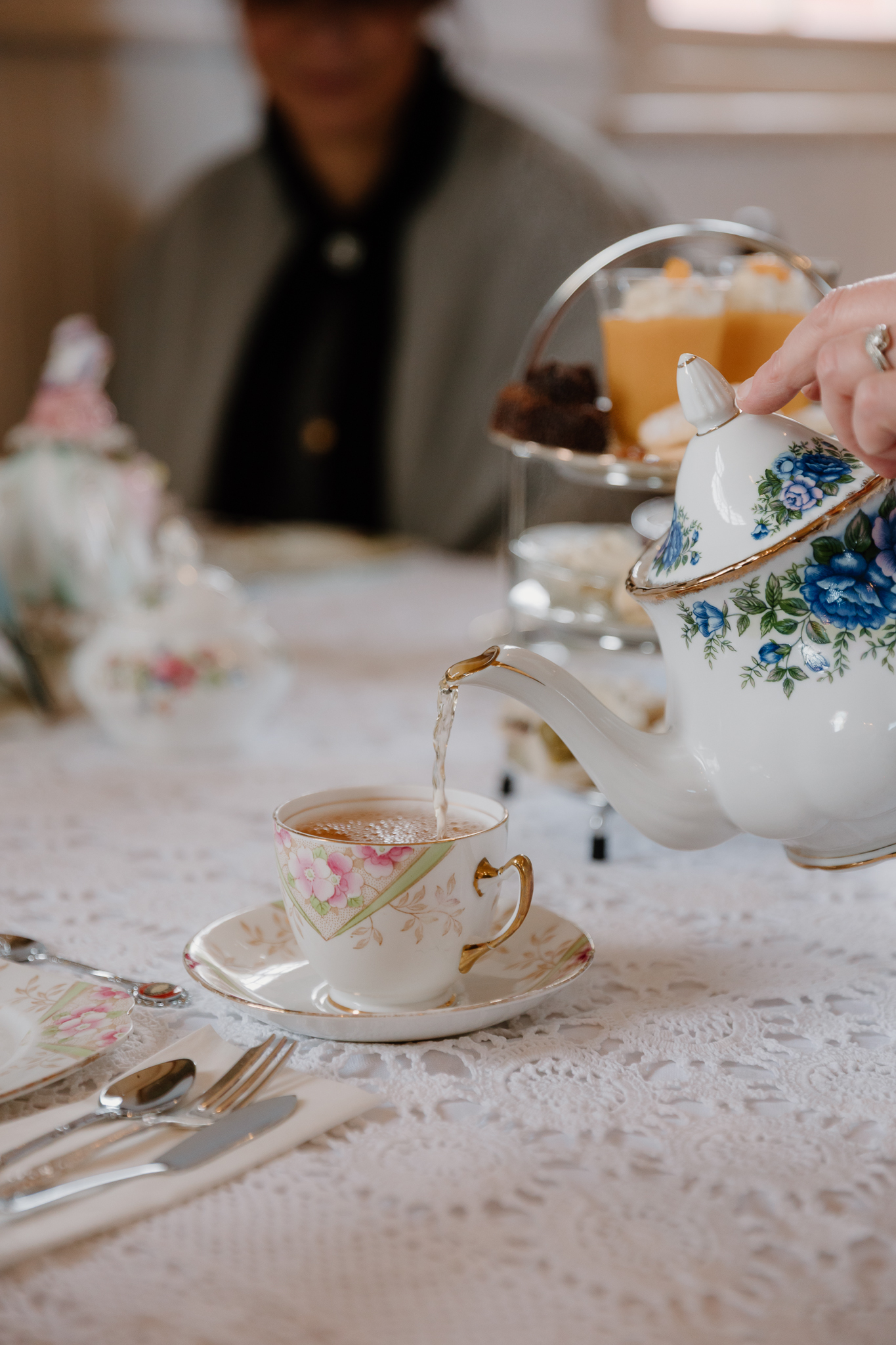 Tea being poured from a floral teapot into a vintage teacup at a lace-covered table, with tiered sweets and a guest in Edwardian-style attire in the background at Willow Bank High Tea.