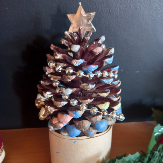 Handmade pine cone Christmas tree decorated with paint, glitter, tinsel and colourful beads on a festive base